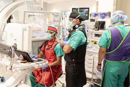 Clinical team members in procedural attire work together in a hospital procedure room with medical equipment and monitors visible.